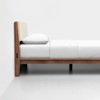 A minimalist wooden bed frame with a light padded headboard, a white pillow, and neatly made white bedding, set against a plain white wall and floor.