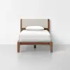 A single wooden bed with a light beige upholstered headboard, white bedding, and a white pillow, set against a plain white wall and floor.
