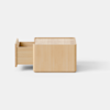 A small, light wooden cube sits on a smooth, light gray surface against a plain white background. The design is minimalistic and modern.