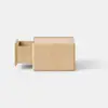 A small, light wooden cube sits on a smooth, light gray surface against a plain white background. The design is minimalistic and modern.