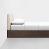 A neatly made bed with a beige upholstered headboard, white pillow, and white bedding, set on a dark wooden frame against a plain white wall and light gray floor.