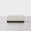 A square, cream-colored ottoman with a textured fabric surface and a dark wooden base, positioned on a light gray floor against a white background.