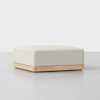 A square, light beige ottoman with a cushioned top and a wooden base sits on a smooth, light gray surface against a plain, light background.