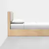 A minimalist wooden bed with a padded headboard, white pillow, and white bedding, positioned against a plain white wall and on a gray floor.