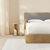 Minimalist bedroom with a wooden bed frame, gray cushioned headboard, white bedding, and a matching wooden nightstand holding books, a glass, and a carafe. Light curtains and a soft area rug complete the calm scene.