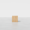 A small, light wood square cabinet with two drawers, placed on a smooth, light gray floor against a plain white background.