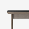 A close-up view of a wooden bench with a rectangular, dark gray cushioned seat, shown against a plain white background. Only part of the bench is visible, focusing on one corner and its leg.