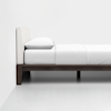 A minimalist bed with a white upholstered headboard, white bedding, and a pillow, set on a dark wooden frame against a plain white wall and floor.
