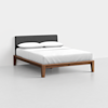 A modern bed with a wooden frame, a dark gray upholstered headboard, a white mattress, and two white pillows, set against a plain light gray wall and floor.