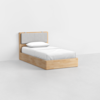 A single wooden bed with a light wood frame, cushioned gray headboard, and white bedding is positioned against a plain white wall and floor in a minimalistic room.