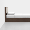 A modern wooden bed with a rectangular beige upholstered headboard, white bedding, and a single pillow, placed against a plain white wall on a light gray floor.