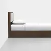 A modern wooden bed with a rectangular beige upholstered headboard, white bedding, and a single pillow, placed against a plain white wall on a light gray floor.