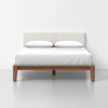 A modern wooden bed frame with a light fabric headboard, made up with white bedding and two pillows, set against a plain white wall and gray floor.