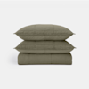 Two olive green pillows are stacked on top of a matching folded quilt, all featuring a subtle stitched grid pattern, against a plain white background.