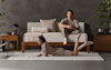 A woman lies on her stomach reading a book on a rug, while a man sits on a white daybed behind her. The room has minimalist decor with neutral tones, wooden furniture, and plants.