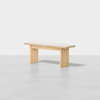 A minimalist wooden bench with a light-colored seat and solid rectangular legs, placed on a plain light gray floor against a white background.