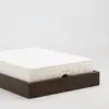 A white mattress with THUMA embroidered on the side sits on a dark wooden bed frame against a plain white background.