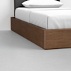 A close-up of a modern bed with a wooden frame, neatly made with white bedding and a pillow, on a light gray floor.