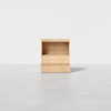 A minimalist wooden chair with a boxy, geometric design and light wood finish, set against a plain white background on a smooth gray floor.