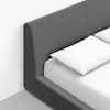 Close-up of a modern bed with a dark gray upholstered headboard and frame, white bedsheet, and white pillows, set against a white wall and light-colored floor.