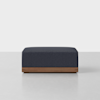 A modern, rectangular ottoman with dark blue upholstery and a wooden base, placed on a light gray floor against a plain white background.