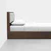 A side view of a modern bed with a dark wooden frame, a cushioned headboard, white bedding, and a white pillow, set against a plain white wall and a light gray floor.