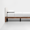 A modern wooden bed frame with a light upholstered headboard, dressed in white bedding and a pillow, placed against a plain white wall in a minimalistic room.