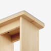 Close-up view of the corner of a light wood table with a slightly rounded edge and a gray cushioned top, showing smooth craftsmanship and clean lines against a white background.