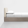 A minimalist bed with a light wooden frame, beige upholstered headboard, and white bedding, including a pillow and neatly tucked sheets, positioned against a plain white wall on a light gray floor.