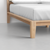 A close-up view of a bed corner, showing a light wooden bed frame with simple, clean lines and a white mattress on top, set on a smooth light gray floor.