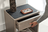 A person’s hand opens a drawer in a light wood nightstand. On top, there is a dark marble vase and a brown tray holding a pair of eyeglasses. Soft daylight enters from a nearby window.