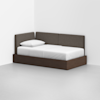 A modern daybed with a dark wooden frame and brown upholstered backrest sits in the corner of a minimalist white room, topped with a white mattress and pillow.