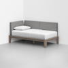 A minimalist daybed with a light wooden frame, white mattress and pillow, and gray upholstered backrest panels, placed in the corner of a bright, empty room.