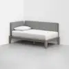A minimalist daybed with a light wooden frame, white mattress and pillow, and gray upholstered backrest panels, placed in the corner of a bright, empty room.