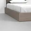 A close-up of a bed with a simple wooden platform frame and white bedding, set on a light grey floor against a plain white wall.