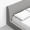 Close-up of a modern bed with a light gray upholstered headboard and frame, featuring a neatly made white mattress and two white pillows. The bed is positioned against a white wall and light gray floor.
