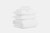 A neatly stacked set of white bedding, including two pillows, a folded comforter, and folded sheets, arranged on a plain white background.
