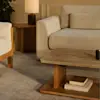 A cozy living room with a beige sofa, a wooden coffee table holding books and a candle, a side table with a lamp, and a textured carpet. Warm lighting enhances the neutral and minimalist decor.
