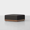 A modern, square ottoman with a dark charcoal upholstered cushion and a wooden base, set against a plain white background.