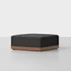 A modern, square ottoman with a dark charcoal upholstered cushion and a wooden base, set against a plain white background.