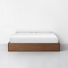 A minimalist bed with a wooden base and white bedding, including two pillows, is placed against a plain white wall on a light gray floor.