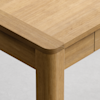 Close-up of the corner of a wooden table with rounded edges and legs, showing the smooth, natural grain of the light wood against a light gray background.