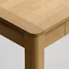 Close-up of the corner of a wooden table with rounded edges and legs, showing the smooth, natural grain of the light wood against a light gray background.