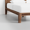 A close-up view of a wooden bed frame with clean lines and a white mattress and pillow, set on a light gray floor against a white wall.
