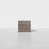 A wooden chest of drawers with a minimalist design, featuring three horizontal drawers, placed on a light gray floor against a plain white background.