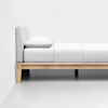 A minimalist wooden bed frame with a light gray upholstered headboard, dressed in white bedding and a single white pillow, positioned against a plain white wall and floor.