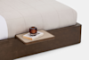 A close-up of a bed with a beige quilt, showing a built-in wooden nightstand holding an amber glass candle and two books. The background is white.