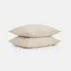 Two plain, light beige pillows are stacked on top of each other against a white background.