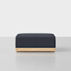 A modern, rectangular ottoman with a dark upholstered cushion and a light wooden base, placed on a neutral surface with a plain white background.
