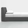 A modern, minimalist bed with a dark gray upholstered frame and headboard, neatly made with white bed sheets and a white pillow, set against a plain white background.